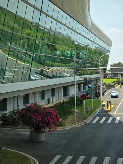 The image shows the exterior of a modern building with a curved glass facade. The glass reflects a lush green hillside with trees and some sky. In the foreground, a road curves around a large planter filled with vibrant pink flowers. A white car with a blue stripe drives on the road, approaching a yellow barrier. Several streetlights stand along the roadside. In the background, a construction vehicle with a raised platform is parked. The scene appears to be a public space, possibly an entrance to a complex or facility, under a bright, partly cloudy sky. The architecture suggests a contemporary urban environment.