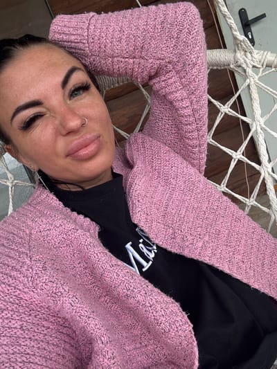 A woman is lying on a hammock, wearing a pink knitted cardigan and a black t-shirt. She is looking at the camera with one eye closed. The hammock is made of white rope and is suspended from a white pole. The background shows wooden panels and a door handle. The image is taken in a close-up shot, focusing on the woman's face and upper body. It is not possible to determine the specific location in Северянин Снт, Russia from the image.