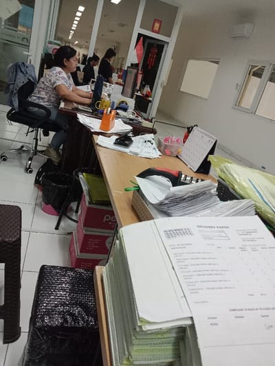 The image depicts an indoor office scene in Semarang, Indonesia, dated October 13, 2023, within what appears to be a garment-related business. A woman with dark hair, wearing a floral top and blue jeans, is seated at a black office chair in the foreground, actively typing or operating a mouse at a wooden desk. A denim jacket is draped over her chair. The desk is cluttered with office supplies including a blue water bottle, a yellow mug, an orange tape dispenser, a calculator, a hole punch, and stacks of papers.

In the immediate foreground, a significant stack of "DELIVERY PAPER" documents is visible, with the top sheet explicitly detailing "Semarang, 13-10-2023" and "GARMENT #1" along with itemized particulars. Underneath the desk, a black trash bin with a pink base, several stacked pink cardboard boxes, and a black plastic-wrapped item are visible on a white tiled floor.

In the background, two additional women are seated at desks, engaged in similar work. The office features white walls, an overhead air conditioning unit, and two frosted or reflective windows. A doorway with a red frame displays a red banner and a red decorative hanging, possibly a Chinese knot or lantern, indicating cultural decor. Fluorescent lighting is visible on the ceiling in the background.