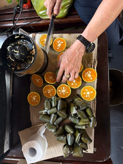 The image, taken from a high-angle perspective, shows a person preparing citrus fruit, possibly oranges, in what appears to be a domestic setting, potentially in the city of Huế, Vietnam. The person, wearing a black watch and a gold ring, is using a knife to cut the oranges in half on a wooden surface, likely a cutting board or table. Several orange halves are already prepared and arranged on the board. A black citrus juicer is positioned near the sliced fruit, suggesting the oranges are being prepared for juicing. Adjacent to the oranges, a stack of leafy green packages, possibly wrapped food items, are present. A roll of white paper towels rests near the food packages. The background shows parts of furniture and possible food packaging, providing further context to the domestic setting.