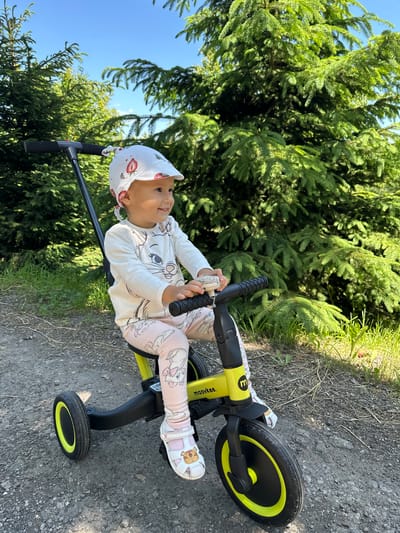 A young child, wearing a white hat with a strawberry pattern, a long-sleeved white shirt with a cat graphic, and pink leggings with cartoon drawings, sits on a bright yellow and black tricycle. The child is smiling and appears to be holding onto the handlebars. The tricycle is on a gravel path. In the background, there are tall green evergreen trees and a bright blue sky. The scene suggests an outdoor setting, possibly a park or a wooded area. The image appears to have been taken on a sunny day. The location is not specified as Wisła, Poland, in the provided image.