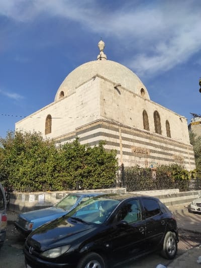 The image depicts a street scene in Al-Qanawat Municipality, Syria, dominated by a large stone building under a partially cloudy blue sky. The central structure features a prominent, light-colored dome topped with a decorative finial. Its facade is constructed from light-toned stone blocks, with horizontal sections of striped ablaq masonry visible on the lower levels. Multiple arched windows with light-colored frames are present on the building's upper walls.

In the foreground, a paved street runs horizontally, with several vehicles parked along it. A black two-door hatchback, resembling a Peugeot 206, is prominently positioned in the lower-center, facing right. Behind it and to the left, a light blue sedan is parked. The rear portion of a silver or grey vehicle is partially visible on the far left. To the far right, a white sedan is partially visible further down the street.

Separating the street and the main building is a low wall or planter, partially adorned with affixed papers or posters, topped by a dark metal fence. Behind this fence and wall, dense green foliage, consistent with bushes or small trees, lines the base of the building. No individuals are visible in the scene.