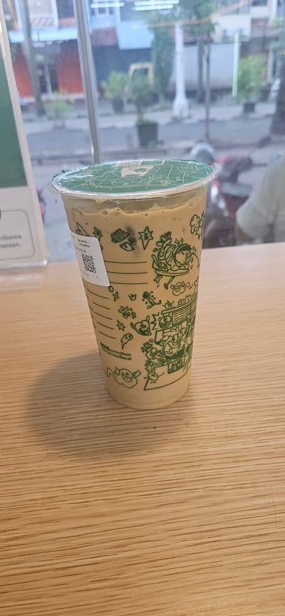 A clear plastic cup filled with a light brown beverage, likely a coffee drink, sits on a light-colored wooden table. The cup is decorated with a green graphic pattern of cartoon characters and small illustrations, giving it a playful aesthetic. A clear plastic lid seals the top of the cup, with some condensation visible on the inside. A small white label with a QR code is affixed to the side of the cup.
The background is blurred, but appears to be a window looking out onto an urban scene. Buildings with windows and some greenery are visible, suggesting an indoor setting with a view of the outdoors. The lighting suggests it could be daytime.
The overall impression is a casual, everyday scene, perhaps in a cafe or a home. The focus is on the beverage in the cup, with the surrounding environment providing context. There is no explicit indication of specific activity beyond the presence of the drink, suggesting it's ready to be consumed.