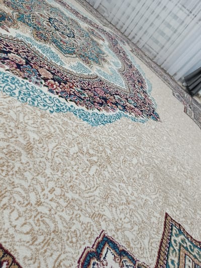 The image shows a close-up, angled view of a Persian-style rug with intricate patterns. The rug has a cream-colored background with a repeating, detailed paisley or floral design in shades of beige and tan. Along the top section of the visible rug, there are borders of more complex patterns featuring rich colors like deep red, navy blue, and teal, with delicate floral motifs. The rug is lying on a floor, and in the upper right corner, sheer white curtains are visible, suggesting an indoor setting, possibly a room in Yzgant, Turkmenistan. The lighting appears natural, coming from the direction of the curtains, casting soft shadows and highlighting the texture of the rug's fibers. There are no people or animals present in the image.