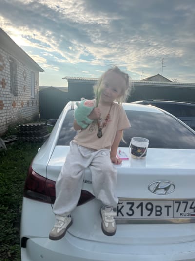 A young girl with blonde hair, wearing a light-colored t-shirt and sweatpants, sits on the trunk of a white Hyundai sedan. She holds a light green plush toy in her left hand and a green stalk in her right. A bucket with yellow items and a small pink object are placed on the trunk next to her. The car's license plate reads "K319BT 774". The background shows a brick building to the left, some stacked grey roofing materials, and a dark green fence. The sky above is partly cloudy. The scene is set in Mal'tsevo, Russia.