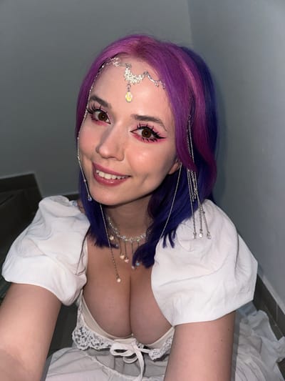 The image shows a woman with purple and blue hair smiling and looking into the camera. She has dramatic pink eyeliner and a decorative silver headpiece. She is wearing a white peasant-style top with a ruffled neckline. The background consists of a gray wall and a small section of what appears to be a floor. No details suggest the scene is located in the City of Zagreb, Croatia.