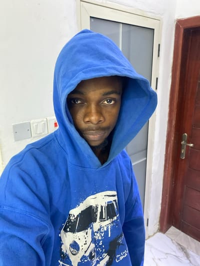 The image features a dark-skinned man wearing a blue hooded sweatshirt (hoodie), looking directly into the camera. He has a beard or stubble, and the hood is up, framing his face. The blue hoodie has a prominent graphic design on the right chest and shoulder area, rendered in white and black. This design incorporates elements resembling the front of a vehicle (possibly a truck or car), a basketball, and paint splatters. Above these, the text "On The LAND" is clearly visible.
The background consists of a plain white wall. To the man's left, a white-framed window is present, allowing natural light into the space. Above the window, there is a dark wooden structure, possibly a door frame or an overhead beam, with a metallic latch visible. To the man's right, on the white wall, a set of white and gray electrical switches or outlets can be seen. The setting appears to be an indoor environment. The man maintains a calm, neutral expression. The image is oriented 90 degrees clockwise from what would be a standard upright portrait. The location context is Joma, Ghana.