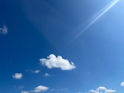 The image shows a bright blue sky with scattered white cumulus clouds. A strong ray of sunlight is visible in the upper right corner of the frame. A contrail of an airplane is visible in the lower right corner. No people or objects are discernible that would indicate the image was taken in Chambœuf, France. The scene primarily consists of atmospheric elements.