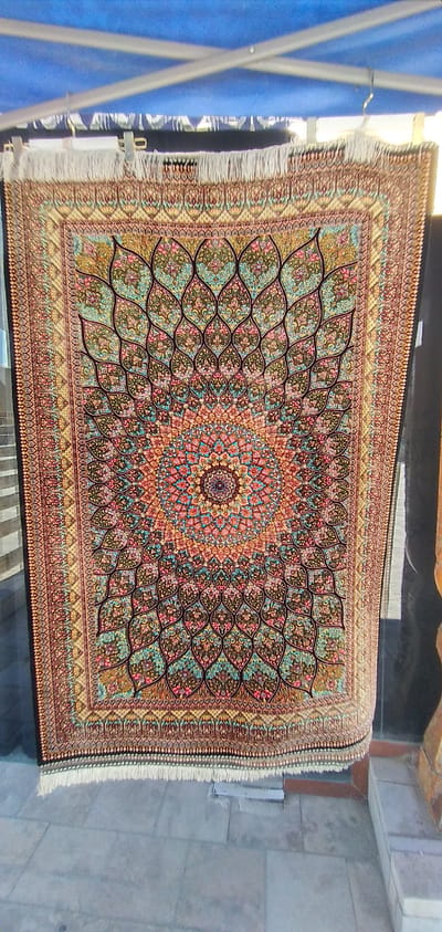 A hanging Persian rug with intricate floral patterns is displayed outdoors, possibly for sale, in Bukhara, Uzbekistan. The rug features a central medallion with vibrant colors including red, orange, blue, and yellow, surrounded by repeating petal-like designs in shades of green, teal, and pink, all set against a dark background. The rug has a decorative border with smaller floral motifs and a fringe at the bottom. The setting appears to be a public area with a tiled floor and a stone wall to the right. A blue tarp is visible above, suggesting an awning or temporary shelter. The overall scene is well-lit, indicating daylight.