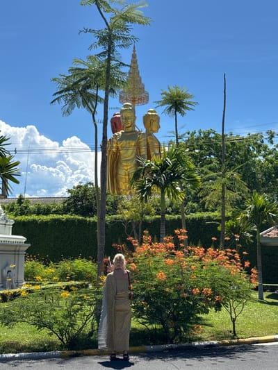 The image shows a landscape in the Bang Pu Subdistrict Municipality, Thailand. Prominently featured is a golden statue of Buddha standing amidst lush greenery, including palm trees and flowering shrubs bearing orange blossoms. A woman is standing near the flora, looking at her phone. A road runs alongside the garden area. The sky is blue with white clouds.