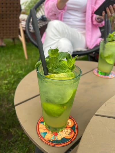 Two glasses of mojitos sit on a table outdoors, with the Vysoké Tatry mountains visible in the background. The glasses are tall and filled with a green liquid, ice, mint leaves, and lime slices. One glass has a black straw. Both glasses are on coasters; one coaster has a stained-glass design of two skeleton figures. A person, wearing a pink jacket and white pants, is partially visible in the upper left of the frame, holding a black tablet. The scene appears to be set in a cafe or restaurant, likely during the daytime, as indicated by the lighting and the presence of outdoor seating and greenery. The overall mood is relaxed and casual.