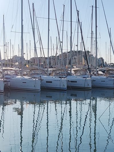 The image presents a serene scene of a marina in Dénia, Spain, filled with numerous sailboats docked side-by-side under a clear, bright blue sky. The foreground and midground are dominated by the sleek, white hulls of modern cruising yachts, their tall masts and intricate rigging creating a dense, vertical network against the sky.
The water in the marina is exceptionally calm, perfectly reflecting the masts and rigging in shimmering, elongated patterns, adding a dynamic yet tranquil quality to the lower half of the frame. Many of the boats feature light-colored canvas covers over their cockpits.
In the background, a hillside is visible, densely covered with multi-story buildings, typical of a vibrant Mediterranean coastal town. The bright daylight suggests it is either morning or late afternoon, with clear weather and gentle illumination. No people are visible, contributing to the peaceful atmosphere.
Visible text on the boat hulls includes the name "Oceanis Coralia" and various registration numbers such as "6ª AT-3-27-22", "6ª AT-3-31-19", and "6ª AT-3-9-10". A small Spanish flag can also be faintly observed flying from one of the distant boats.