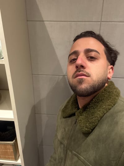 A man poses indoors, partially visible and rotated 90 degrees counter-clockwise. He has dark hair, a beard, and brown eyes, wearing a green suede jacket with a fur collar. The background features grey tiled walls and a white shelving unit with visible items. The lighting creates shadows on the wall.