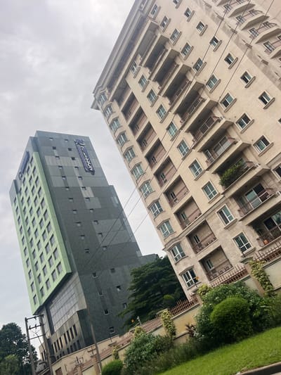 In the city of Ikoyi, Nigeria, this image captures a view of two modern buildings against an overcast sky. The building on the left is a tall, slender structure with a facade of alternating light green and dark gray panels. Several small, dark square windows are visible on the dark gray sections, while a grid of rectangular windows covers the green sections. A prominent banner with the word "RADISSON" in white lettering on a dark blue background is affixed vertically to the side of this building. The building on the right is a beige, multi-story residential or office building with a repeating pattern of balconies and windows. Each balcony has a brown metal railing. The lower part of the image shows a well-maintained green lawn and various shrubs and trees. A low, ornate brown fence with climbing plants runs along the edge of the property. Electrical poles and wires are also visible in the foreground, indicating urban infrastructure. The perspective is low-angle, emphasizing the height of the buildings.