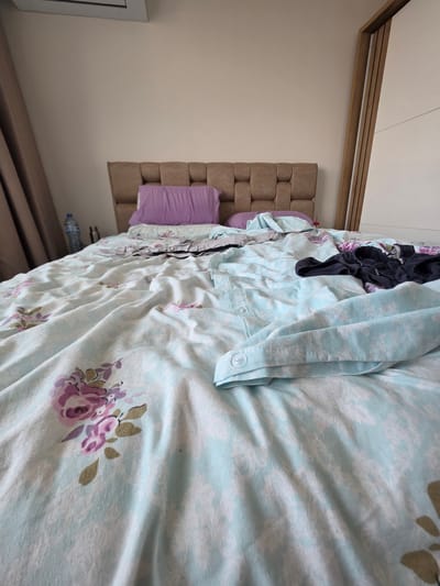 The image shows a messy bedroom, likely in Yagodovo, Bulgaria. The bed is unmade, with blankets and pillows scattered. There are clothes lying on the bed, including a dark t-shirt and a pair of dark shorts. A water bottle is visible on the bedside table. The room has a light-colored wall and a wooden headboard. A large wardrobe with a white door is also present. The overall impression is one of a lived-in and somewhat untidy space.