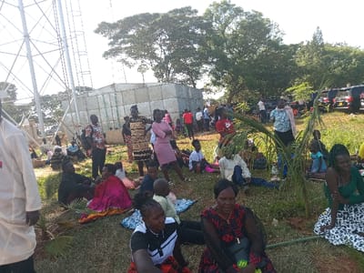 This outdoor image captures a diverse group of people, including adults, children, and infants, gathered on a grassy area in Siaya, Kenya. Many individuals are seated directly on the ground or on small blankets, while others stand and mingle.
In the foreground and midground, people are informally arranged across the scene. Some individuals are looking towards the camera, while others are engaged in conversation or observing their surroundings. A woman in a pink dress stands prominently in the midground, and several children are visible sitting near adults. Small palm fronds are scattered on the ground, possibly indicating a casual or celebratory setting.
The background features several structures. To the left, a tall, white metal lattice structure, possibly a communication or water tower, dominates the skyline. Behind the gathered people, there's a large, cuboid structure, appearing to be made of corrugated metal or panels, which could be a storage tank or building. Further in the background on the right, several dark-colored SUVs or cars are parked, suggesting proximity to a road or parking area. Large green trees frame the upper right portion of the image, and a clear, bright sky indicates sunny weather, likely during midday or early afternoon. No discernible text is visible on any objects or clothing in the image. The overall impression is that of a community gathering in an open-air setting.