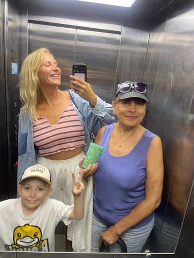 The image shows three people in an elevator. A blonde woman is taking a selfie, smiling broadly. She is wearing a pink and black striped crop top and a denim jacket. Next to her stands an older woman with short brown hair, wearing a purple tank top and a plaid baseball cap with sunglasses on top. She is holding a green cup. To the left of the older woman and in front of the blonde woman is a young boy wearing a white t-shirt with a yellow cartoon character on it and a beige and black baseball cap. He is making a peace sign with his fingers. The elevator interior is metallic with visible scratches and scuff marks. The scene is consistent with an indoor setting like an elevator in Giachrypsh, Abkhazia.