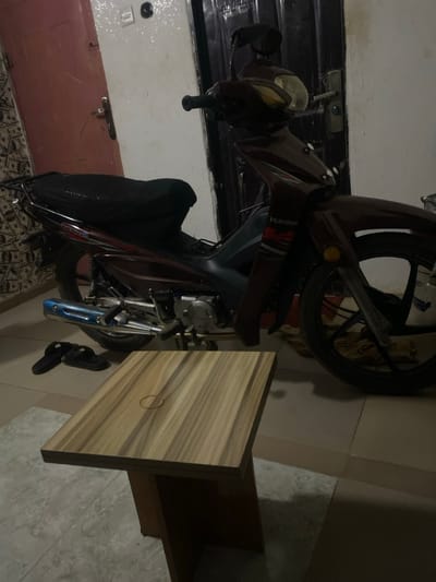 A maroon scooter stands beside a wooden cabinet with a door. The scooter features a black seat, a headlight with a gold tint, and a blue exhaust pipe. One wheel is visible, and what appear to be sandals are situated near the scooter's base, close to the cabinet. The background includes a tiled floor, a door, and parts of a wall. The image is rotated counterclockwise.