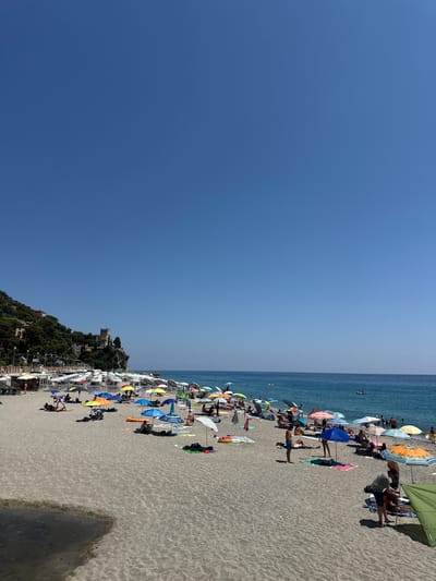 The image depicts a sunny day at the beach in Finale Ligure, Italy, specifically the Spiaggia di Finale Marina. The scene is dominated by a vast expanse of sandy beach, dotted with numerous colorful umbrellas providing shade for beachgoers. The sea is a vibrant blue, with a few small boats visible in the distance.

On the left side of the image, a lush green hillside rises, topped by a historic castle-like structure, which appears to be the Castel Gavone. The hillside is covered with dense vegetation and a few buildings are visible nestled among the trees. Along the edge of the beach, there are rows of beach chairs and umbrellas set up for rent.

People are scattered across the beach, some lounging under umbrellas, others walking or playing in the sand. In the foreground, a small pool of water has formed on the sand, possibly from a recent tide or a small stream. The overall atmosphere is one of leisure and relaxation on a warm summer day.