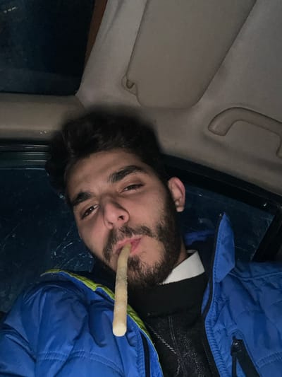 A selfie shows a man with dark hair and a beard, lying on his side, possibly in a car. He has something long, white, and cylindrical in his mouth. He's wearing a blue puffy jacket. The interior of the car is visible, including a sun visor and a grab handle. The image is dimly lit.