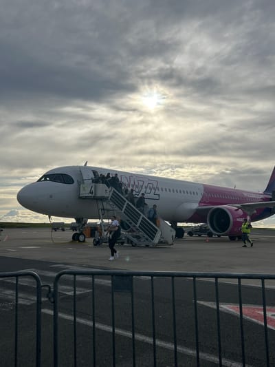 The image displays an active airport scene featuring the boarding of a Wizz Air airplane on a tarmac under a cloudy sky. The aircraft, predominantly white with a distinctive pink/purple tail and engines, is parked with its nose facing the left side of the frame. Passengers are in the process of boarding via mobile stairs positioned at the front door of the aircraft, with several individuals visible on the steps. Ground crew personnel, one in a high-visibility vest, are present near the aircraft's engines and nose area.

The sky is heavily overcast, yet a bright sunburst pierces through the clouds, indicating either early morning or late afternoon. A dark metal fence runs along the right side of the foreground, separating the viewer from the airport apron. The tarmac shows various ground markings and a faded large red marking that is indistinct.

Visible text on the aircraft includes "Wizz Air" along the fuselage and "wizzair.com" near the tail. A sign attached to the mobile boarding stairs clearly displays "Dortmund Airport," confirming the image's location as Dortmund, Germany. Additional text "L 4" is also visible on the stairs. The scene captures the functional aspect of air travel, with no specific emotions discernible from the individuals depicted.