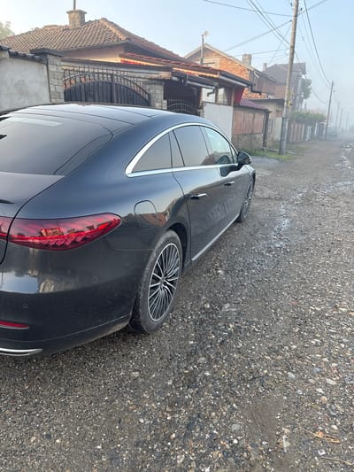 A dark-colored, possibly black or dark grey, modern sedan is parked on an unpaved, gravel road. The car, which appears to be a Mercedes-Benz with sleek lines, multi-spoke alloy wheels, and distinctive LED taillights, is visibly dusty on its bodywork. It occupies the right and central foreground of the image, angled towards the left.
To the left of the car, a series of traditional houses line the street. These buildings feature brick walls, pitched roofs with reddish-brown tiles, and some have ornate, possibly wrought-iron, fencing and an arched gate. Utility poles with a dense network of overhead wires run parallel to the road and houses.
The road surface is composed of loose gravel and dirt, with various sized stones and some areas that appear damp or muddy. The ambient light is soft and diffused, suggesting early morning. A noticeable haze or light fog hangs in the air, particularly in the background, obscuring distant details and contributing to the soft lighting. There are no people visible, and no specific activity is taking place beyond the car being parked. No visible text is clearly discernible in the image. The scene suggests a setting in a village or less developed area, consistent with Kran, Bulgaria.