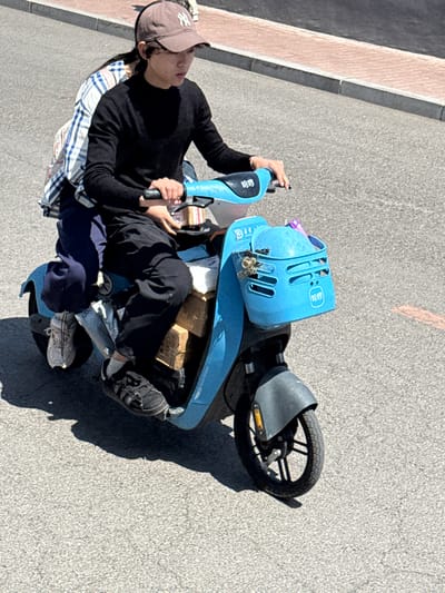 A person sits on a blue electric scooter loaded with packages, with a second scooter on the ground next to it, as viewed from directly above. The scooter is parked on grey asphalt, with a portion of a sidewalk visible on the left edge. The person, wearing a dark long-sleeved shirt, dark pants, and grey sneakers, is seated on the scooter amidst various items, including cardboard boxes and what appear to be packages. They are also wearing a baseball cap with a "NY" logo. A blue basket is affixed to the front of the scooter. The second scooter is lying on the ground next to the first one. The scene is brightly lit, casting shadows of the scooter and person on the pavement. The city of Zhongtun, China is not identifiable.