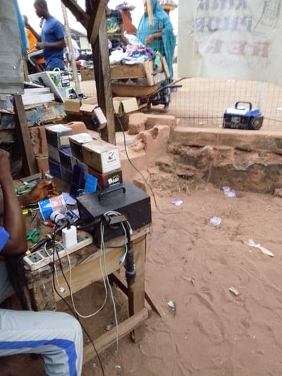 An outdoor market setting features a person partially visible, seated near a wooden workbench. The bench holds an electrical power box, a power strip with various charging cables plugged in, and tools. Stacked boxes, possibly containing goods for sale, are nearby. In the background, a woman in traditional clothing stands near a wheelbarrow filled with merchandise. A blue radio sits on a ledge behind a chain-link fence, and the ground is sandy with scattered debris.