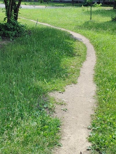The image shows a narrow, winding dirt path in a grassy area. The path appears to be well-worn, suggesting it is a frequently used walkway. Lush green grass lines both sides of the path, with some trees and shadows visible in the background. The scene evokes a sense of a quiet, natural setting within an urban environment. This path is located in the city of Hrodna, Belarus.
