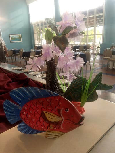 This is a close-up, eye-level, indoor shot of a decorative centerpiece in a dining or event space, possibly a restaurant, located in Pedro González, Venezuela. The centerpiece features a vibrantly painted red and blue wooden fish as the primary subject, positioned in the foreground. The fish has an open mouth and detailed scales painted in red with black accents. Behind the fish, a darker, textured structure supports a cluster of artificial light purple flowers and green foliage. To the left of the centerpiece, a swath of deep red fabric is draped, suggesting a tablecloth or runner. In the background, several round tables covered with dark blue cloths are visible, each set with chairs. A large window with a grid pattern allows natural light to enter the room, illuminating the space and revealing outdoor foliage. On the left wall, a framed landscape painting of a body of water is hung. The flooring appears to be tiled in a mosaic pattern. A person, partially visible on the far left, is wearing a white shirt and dark apron, indicating they may be staff.