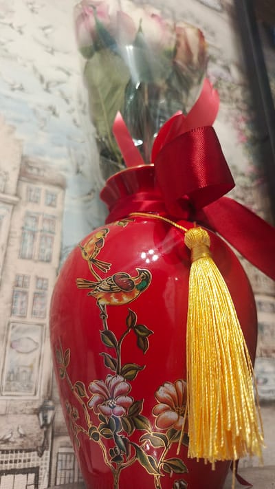 A red ceramic vase adorned with gold-outlined floral and bird motifs is positioned in the foreground. A bright yellow tassel hangs from a string attached to the vase. A red ribbon is tied in a bow around the neck of the vase, with a bouquet of pink roses in cellophane wrapping partially visible behind it. The background features a blurred wallpaper depicting buildings and street lamps, possibly in a European style. The scene is well-lit, with the vase being the primary focus. There is no indication of the city of Zvenigorod, Russia, or any specific location information in the image.