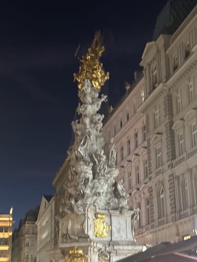 The image depicts the Pestsäule (Plague Column) in Vienna, Austria at night. The column is an ornate baroque monument adorned with sculptures and topped with a gilded pyramid of figures. It is located in the Graben, a prominent pedestrian street in the city center. To the right of the column is a multi-story building with classical architectural features, including arched windows and decorative elements. The sky above is dark blue, indicating it is nighttime. The overall scene is illuminated by artificial lights, casting a warm glow on the monument and surrounding architecture. There are no people clearly visible in the image, and the focus is on the monument and the building.