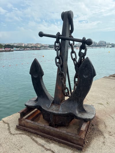 The image depicts a large, dark metal anchor resting on a concrete pier, with a body of water and a city skyline in the background. The anchor is ornate, featuring a stock with two rounded ends and two flukes. Thick, rusty chains connect the anchor to an overhead support structure. The water appears calm and a light blue, with a series of orange buoys visible in the distance. The city skyline features a mix of buildings, including modern high-rises, suggesting an urban coastal environment. The sky is overcast with scattered clouds. The anchor itself is positioned on a rusty metal frame, which is placed on the concrete surface. The concrete shows some cracks and minor debris. The scene is likely in Gelendzhik, Russia, given the context of the city's waterfront and characteristic architecture. There are no people visible in the image. The overall impression is one of a historical maritime artifact displayed in a public space.