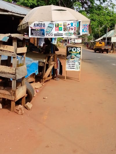 The image displays a roadside commercial establishment in Alor, Nigeria. In the foreground, a multi-tiered wooden shelf structure stands on the left, holding two clear plastic bottles containing yellowish liquid. An old black tire is visible at the base of this structure, resting on reddish-brown unpaved ground, which is scattered with rocks and debris.
Centrally, a large beige octagonal umbrella shades a wooden counter or stall. Attached beneath the umbrella is a banner advertising "AIKITO BUSINESS VENTURES," listing services such as "POS," "Provisions," "Wrappers," "Suits," "Men Slippers," "Women Slippers," "Pepper Soup," "Dry Meat," and "Recharge Card All Network." Images of a POS terminal, mobile network logos, and clothing items are displayed on the banner. A person, partially visible and wearing dark clothing, stands behind the counter.
To the right of the stall, a freestanding A-frame sign with a white board displays "POS AVAILABLE HERE" in prominent black text, alongside images of currency and a POS machine. The sign further lists services: "Cash Deposit," "Withdraw cash," "Send money," "Buy airtime," and "Pay Bills."
An asphalt road extends horizontally across the midground. On the far side of the road, several low-rise buildings with corrugated metal roofs are present. A yellow utility truck with a dark flatbed is parked on the right side of the road. Green tree foliage is visible above the distant structures. Indistinct human figures are walking in the background along the road. The scene is illuminated by daylight, casting soft shadows.