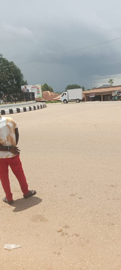 The image depicts a street scene in Kuru Dungus, Nigeria. In the foreground, the lower body of a person wearing a tie-dye t-shirt and red pants is visible, facing away from the camera and walking on an asphalt road. The person is wearing sandals. In the mid-ground, a white refrigerated truck is driving on the road, and a small green tricycle with a rider is parked to the right. Behind the truck, buildings with brown roofs are visible. To the left, a billboard with images of a man and a woman is displayed, and behind it are trees. The road is paved with asphalt and has a curb with black and white stripes. The sky is overcast with dark clouds, suggesting potential rain. A discarded plastic bottle is on the ground in the lower left corner.
