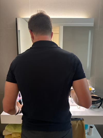 A person is standing with their back to the camera, facing a bathroom sink and mirror. The person is wearing a black polo shirt and dark pants. The bathroom features a modern aesthetic with tiled walls and a lit mirror. On the counter next to the sink are various toiletries and a makeup bag. Below the counter, a stack of folded towels is visible, along with other bathroom supplies. The scene appears to be within a private residence or a hotel bathroom. The city of Murcia, Spain, is not depicted in this image.