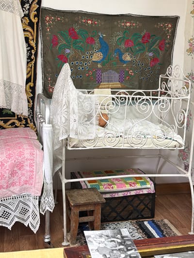 This image depicts an indoor scene that appears to be part of a museum exhibit in Ust'-Pyzas, Russia, showcasing traditional domestic life. The primary focus is a vintage white metal baby crib, ornately decorated with scrollwork. Inside the crib, a doll or baby is swaddled in a patterned blanket. The crib is draped with a delicate white lace cloth.
Behind the crib, a large tapestry hangs on the wall, featuring an embroidered design of peacocks amidst floral motifs in shades of green, blue, red, and purple. To the left of the tapestry, a white fabric with lace trim is visible, along with another patterned textile.
In front of the crib, a small wooden stool stands next to a dark wooden chest topped with a colorful quilted fabric. A small, woven rug is placed on the wooden floor in front of the chest.
To the left of the crib, a bed is partially visible, covered with a pink textile featuring a delicate white pattern, and adorned with white lace trim at its base. The overall impression is one of a preserved domestic setting, likely intended to evoke a sense of nostalgia and cultural heritage.