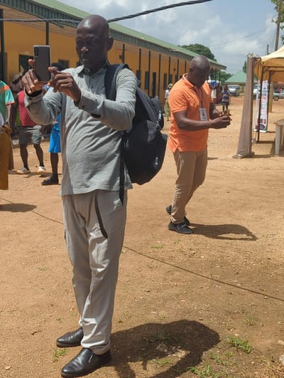 The image shows two men standing on a dirt ground in Maigizo, Nigeria. One man is wearing a grey shirt, light grey pants, and black shoes. He has a black backpack and is holding a black smartphone, seemingly taking a picture. The other man is wearing an orange shirt, beige pants, and black sneakers. He is also holding a smartphone and appearing to take a picture. The background includes a building with a green roof and yellow walls. Other people are visible in the background. The lighting suggests it is daytime. A temporary tent structure with signs is present. The ground is primarily dirt with sparse vegetation.