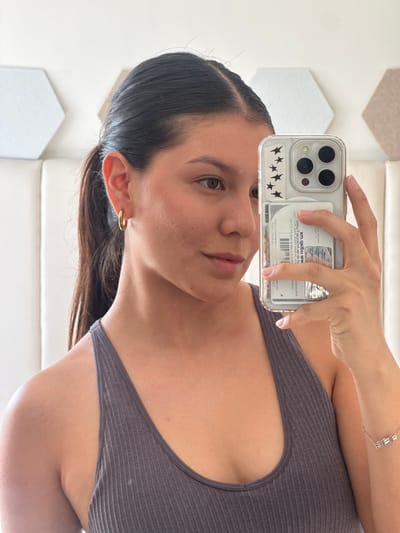 A woman is taking a selfie with her phone. The phone has a clear case with star stickers and a card inserted in the back. The woman has dark hair pulled back, a gold hoop earring, and a grey top. She is wearing a bracelet on her right wrist. The background includes geometric shapes and a white wall. The card visible in her phone case contains the text "xm.qslbu.ww" and other printed details in Spanish. Based on the partial text of "San Francisco Ocotlán" on the card visible in the phone case, the card may be related to this location in Ocotlán, Mexico.