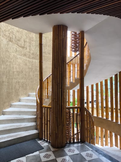 The image captures a spiral staircase in Sidemen, Indonesia. The staircase, constructed of concrete, curves around a central bamboo column, featuring a bamboo railing. The walls surrounding the staircase are textured. Light enters the space through bamboo screens, casting vertical shadows. The floor at the base of the staircase is tiled in a black and white checkered pattern. A gray mat is visible at the bottom of the stairs. The ceiling above is composed of exposed wooden slats.