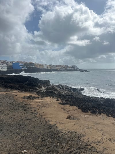 The scene depicts a rugged coastal landscape in San Miguel de Abona, Spain. The foreground is composed of dark, jagged volcanic rock formations intermingled with areas of lighter brown, gravelly soil; minimal debris is visible on the ground in the lower-left. The mid-ground features a turbulent, dark blue-grey sea with white waves breaking against the rocky coastline. In the background, a coastal town is constructed directly on dark cliffs. Buildings within the town display varied colors, including white, yellow, orange, and a prominent bright blue structure on the left side of the town cluster. A small building and a navigational beacon are discernible on a rocky promontory extending into the sea on the right side of the town. The sky is largely overcast with extensive grey and white cumulus clouds, with some patches of lighter blue visible. No individuals are present within the frame.