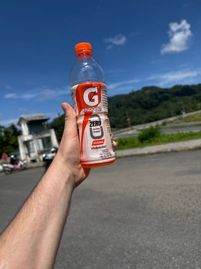 A Caucasian male is holding a bottle of Gatorade Zero. The bottle is orange and white with the Gatorade logo. The background is a blurry image of a road and a hillside covered in lush green trees. There is a small white building with a tan roof in the distance. The sky is a clear blue with a few white clouds. The man's arm is slightly sweaty, indicating a warm climate. The location is the city of Kathu, Thailand.
