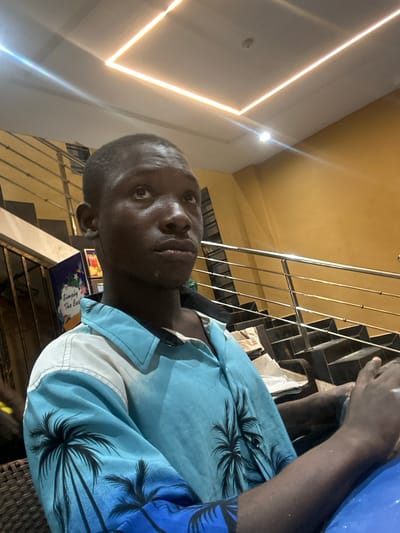 The image shows a young Black male sitting indoors, looking upwards and to the right. He is wearing a light blue short-sleeved collared shirt with a palm tree print. The background features a staircase with a silver railing and a yellow wall. The ceiling has a square recessed lighting fixture with illuminated strips. A sign with text and images is visible on the left side of the frame, near the stairs. The lighting suggests an indoor setting, possibly a restaurant or a public building. Ihiagwa, Nigeria, is a town in Imo State, known for its developing urban infrastructure.