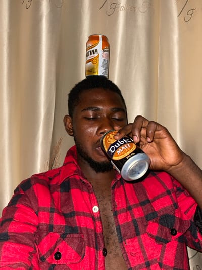 A man in a red and black plaid shirt reclines against a silky gold fabric. He has a beer can balanced on his forehead and another can pressed to his lips, seemingly drinking from it. The beer can on his forehead is orange, while the one he's drinking from is black with gold lettering. His eyes are closed, and he has a dark beard. The scene is casually staged and appears playful.
