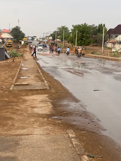 The image depicts a street scene in Minna, Nigeria, on a wet day. The road surface is slick with water. A line of vehicles, including motorcycles, cars, and buses, stretches into the distance. A pedestrian is walking along the sidewalk. Buildings and trees line the street. The sky is overcast.