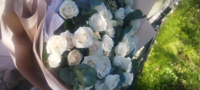 A bouquet of white roses is displayed, with some greenery interspersed. The bouquet is partially wrapped in light-colored paper, which appears to be a soft pink or beige. The roses are in various stages of bloom, with some fully open and others still budding. The bouquet is positioned in what appears to be an outdoor setting, as suggested by the presence of green grass and some small yellow flowers in the background. The lighting indicates it is daytime. No individuals are visible in the image. The context of Yakutsk, Russia, does not directly inform the visual elements of the image itself, as the scene is generic and could be found in many locations.