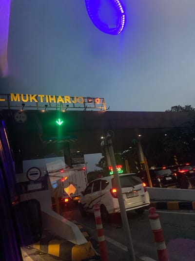 This is a photograph of a toll gate in Semarang, Indonesia, taken at dusk or early evening, as indicated by the dim lighting and the bright artificial lights of the toll plaza. The camera is positioned inside a vehicle, looking out towards the toll booths.

The most prominent subject is the toll gate structure itself, with large, illuminated yellow lettering spelling out "MUKTIHARJOLLO" vertically. A green arrow signal indicates that one of the lanes is open. There are also signs visible, including one that reads "TINGGI MAKS 4.20m" (Maximum height 4.20m) and another with a circular "GERBANG TOL" (Toll Gate) symbol. Traffic lights are visible, displaying red lights in some lanes, suggesting other lanes are closed or occupied.

Several vehicles are present, including a white car and a truck, waiting to pass through the toll gate. The bollards and barriers of the toll plaza are clearly visible, with red and white striped poles and yellow and black striped warning signs. In the upper left of the image, a bright blue, circular light fixture is visible, likely part of the toll plaza's lighting or decoration.

The overall atmosphere is that of a typical evening scene at a busy transportation point. The sky is a muted gray, suggesting it's either overcast or the light is fading. The image captures the functional aspect of a toll collection point with the presence of vehicles, signs, and the toll gate infrastructure.