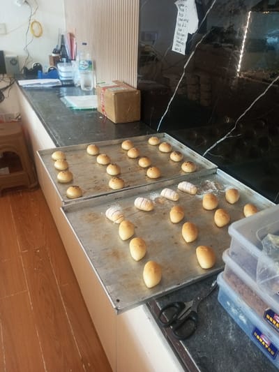 Two trays of baked goods sit on a counter. The top tray holds round buns, some topped with seeds. The bottom tray contains oval buns and rolls with a sugary glaze. A pair of scissors rests near a stack of plastic containers labeled "Donatsu". Behind the trays are a cardboard box, a bottle of water, and other items on the counter. The background includes a wall with a dark, marbled pattern and a textured wall panel. Wooden flooring is visible in the foreground.