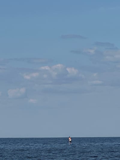 A lone person stands on a paddleboard in the dark blue water of the sea, facing away from the camera. The person is wearing a white and orange swimsuit and is holding a paddle. The sea is calm with small waves. The sky is a clear light blue with some scattered white clouds. The horizon is visible in the distance, separating the sea from the sky. The image appears to be taken during the daytime. The scene does not show any specific landmarks or indications of the location in Demydiv, Ukraine.