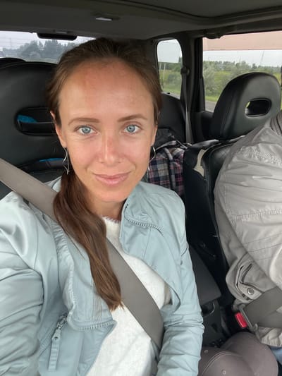 A woman with long brown hair and blue eyes is sitting in the passenger seat of a car. She is wearing a light blue jacket over a white sweater and a grey seatbelt. She is looking at the camera and smiling. The interior of the car is visible, including black leather seats. Outside the car, a green landscape with trees and a cloudy sky can be seen through the window. The location is Petrovskii, Russia.